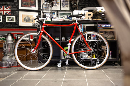 Vélo fixie Abici blanc avec cadre en acier et guidon droit, posé sur une surface en bois.